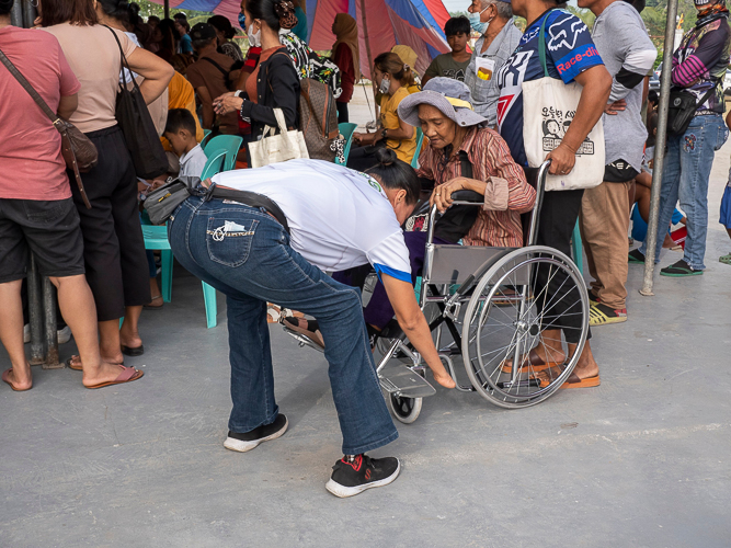 Maria Fe “Nene” Socias (in white) dutifully assists a senior citizen settle into a wheelchair while waiting for her turn at the medical mission. 【Photo by Matt Serrano】