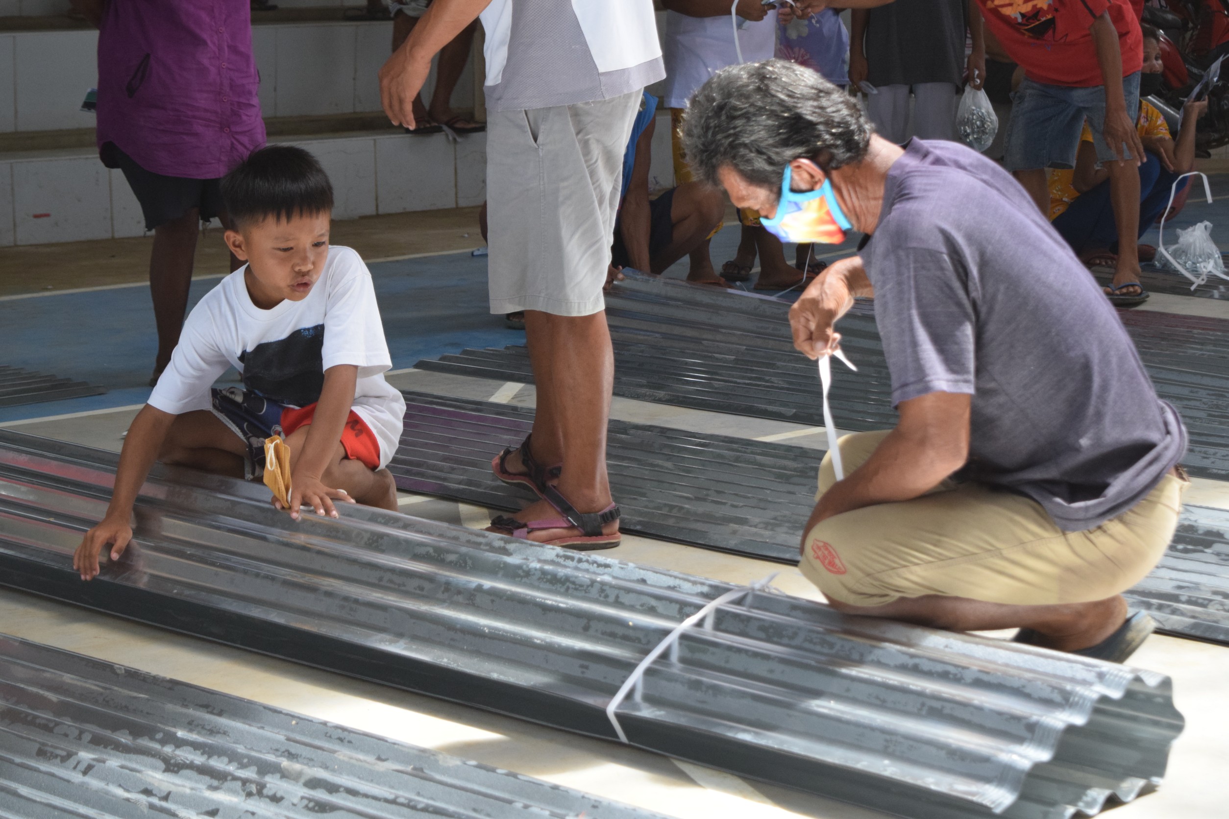 A boy holds down a folded GI sheet as a man ties it down for easier transport. 