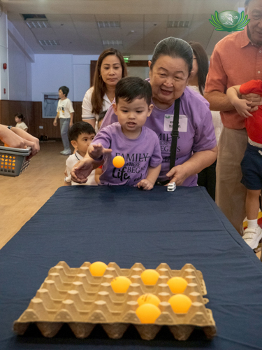 A grandmother happily encourages her grandson to play the egg carton pingpong ball game.