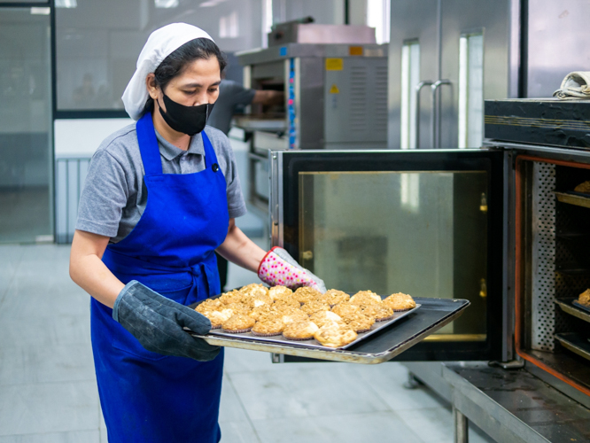 A kitchen volunteer loads a pan of banana streusel cupcakes into an oven. 【Photo by Daniel Lazar】