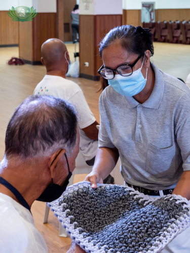 A volunteer shows a beneficiary a sample of the upcycled floor mat woven out of loop lines or excess material from sports socks.【Photo by Daniel Lazar】