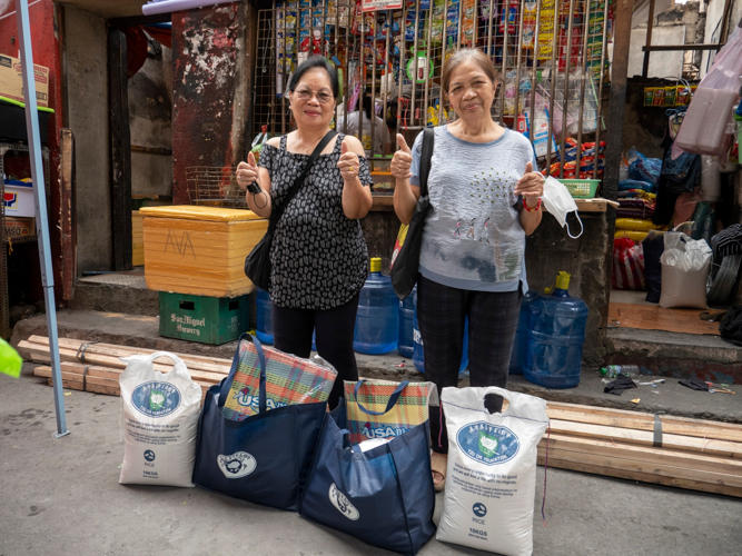 With neighbor Delia Lazarra (left), Lulu Osit signs “thank you” to Tzu Chi for its relief operations. Behind them is Lulu’s store, which burned down, save for its façade. Since the fire, she has used the store front to restart her small business. 【Photo by Matt Serrano】